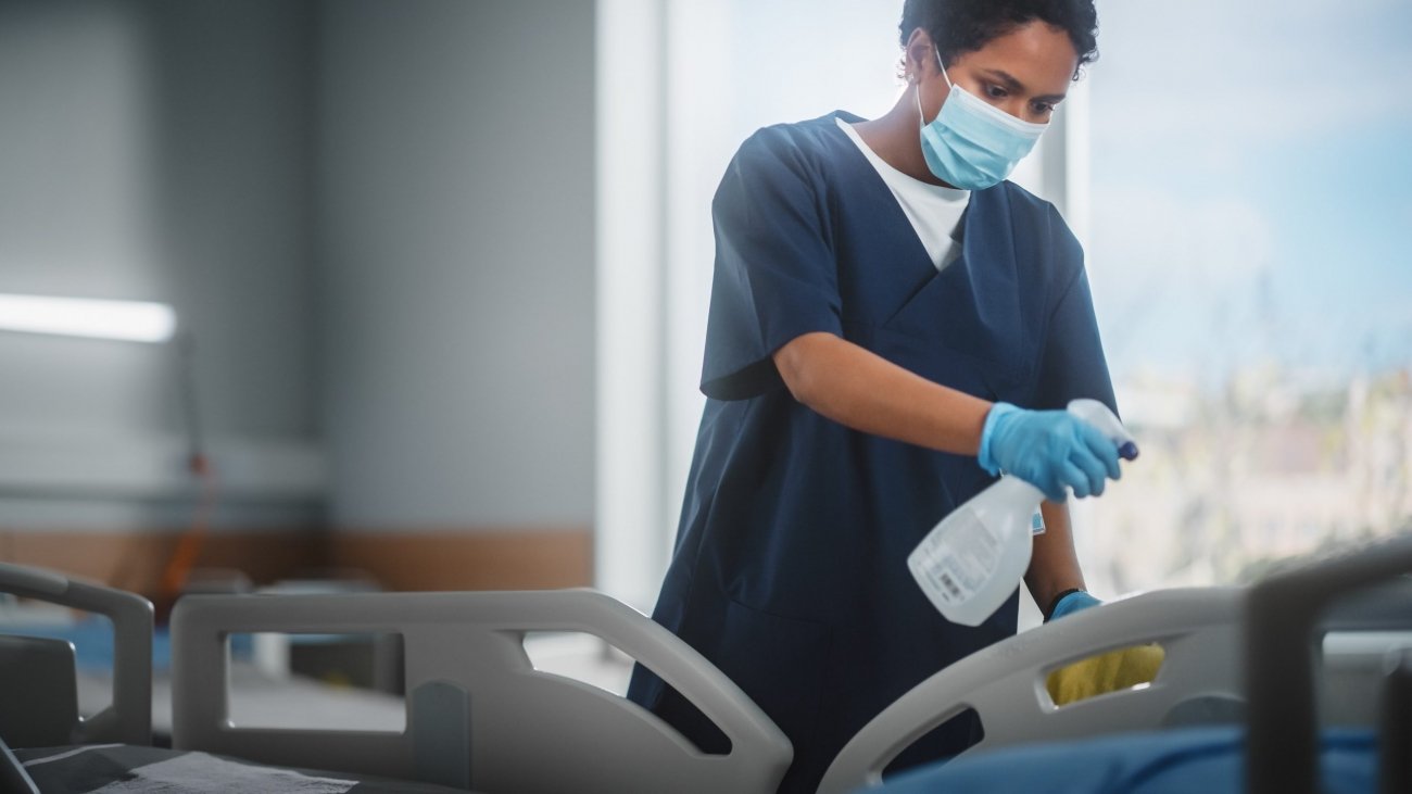 Hospital Ward: Professional Black Nurse Wearing Face Mask, Wiping the Bed, Cleaning Room After Covid-19 Patients Recover. Disinfection, Sterilizing, Sanitizing Clinic after Coronavirus Infected People